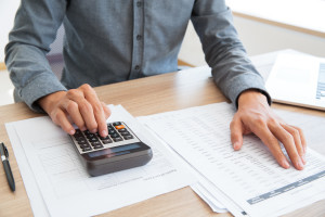 Close-up of unrecognizable accountant calculating company income and examining report. He using calculator to check financial data. Man working at table. Investment concept